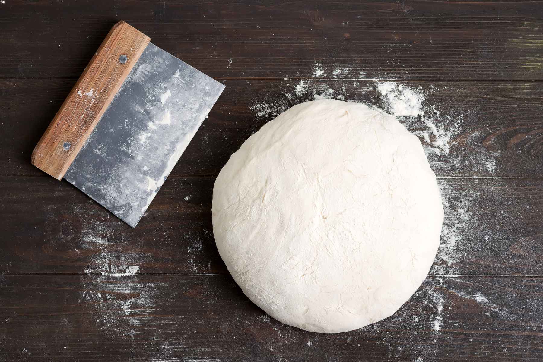 Bench scraper for baking next to batch of bread dough on wooden tabletop. Favorite kitchen tools.