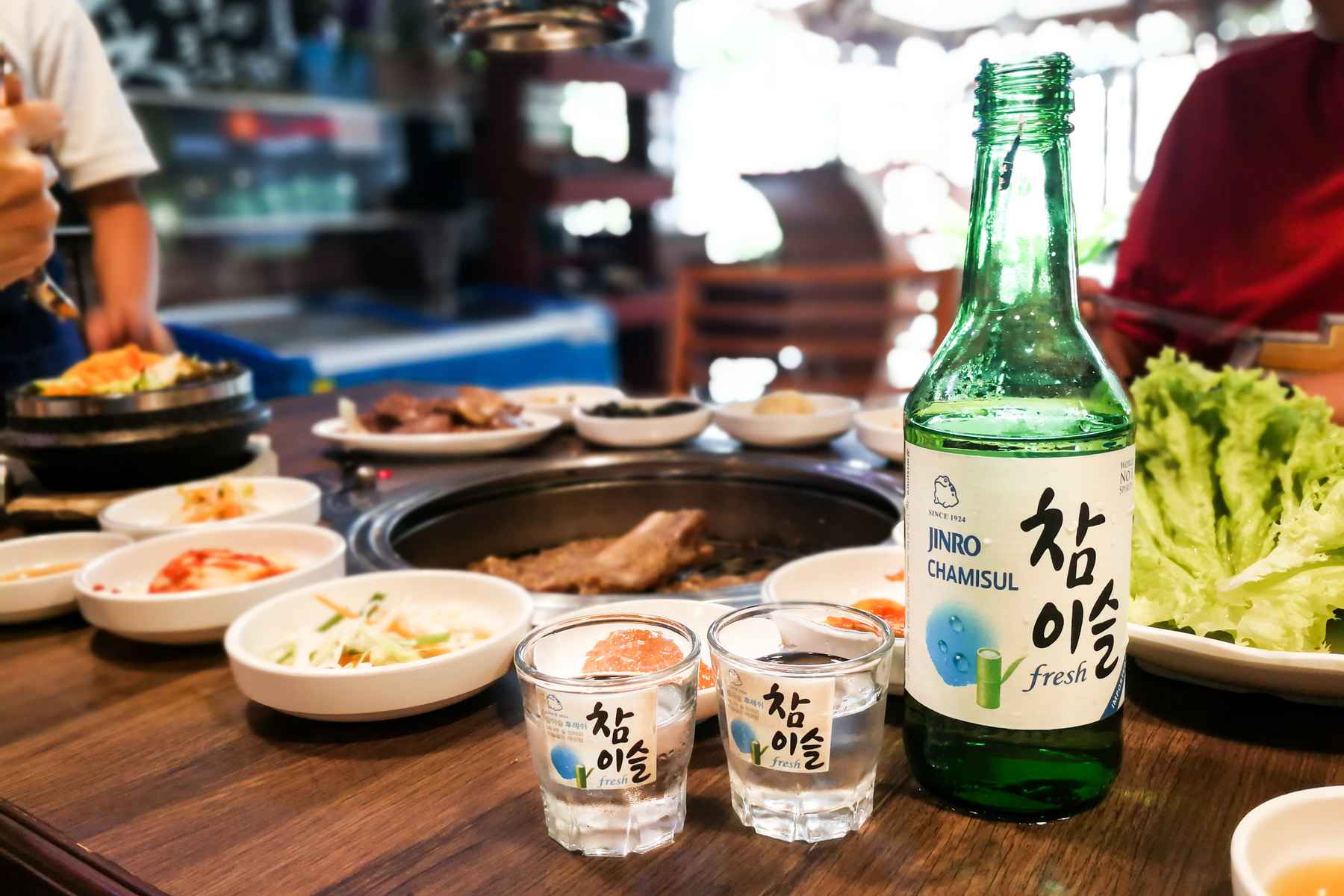 Bottle of soju and two shot glasses on a restaurant table next to a spread of Malaysian food.