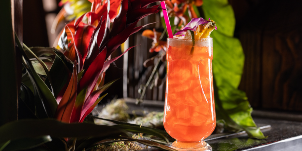 A vibrant orange-pink tropical cocktail from Adrift Tiki Bar in Denver is garnished with fresh fruit and a purple orchid. It has a neon pink straw and is set against a backdrop of tropical flora