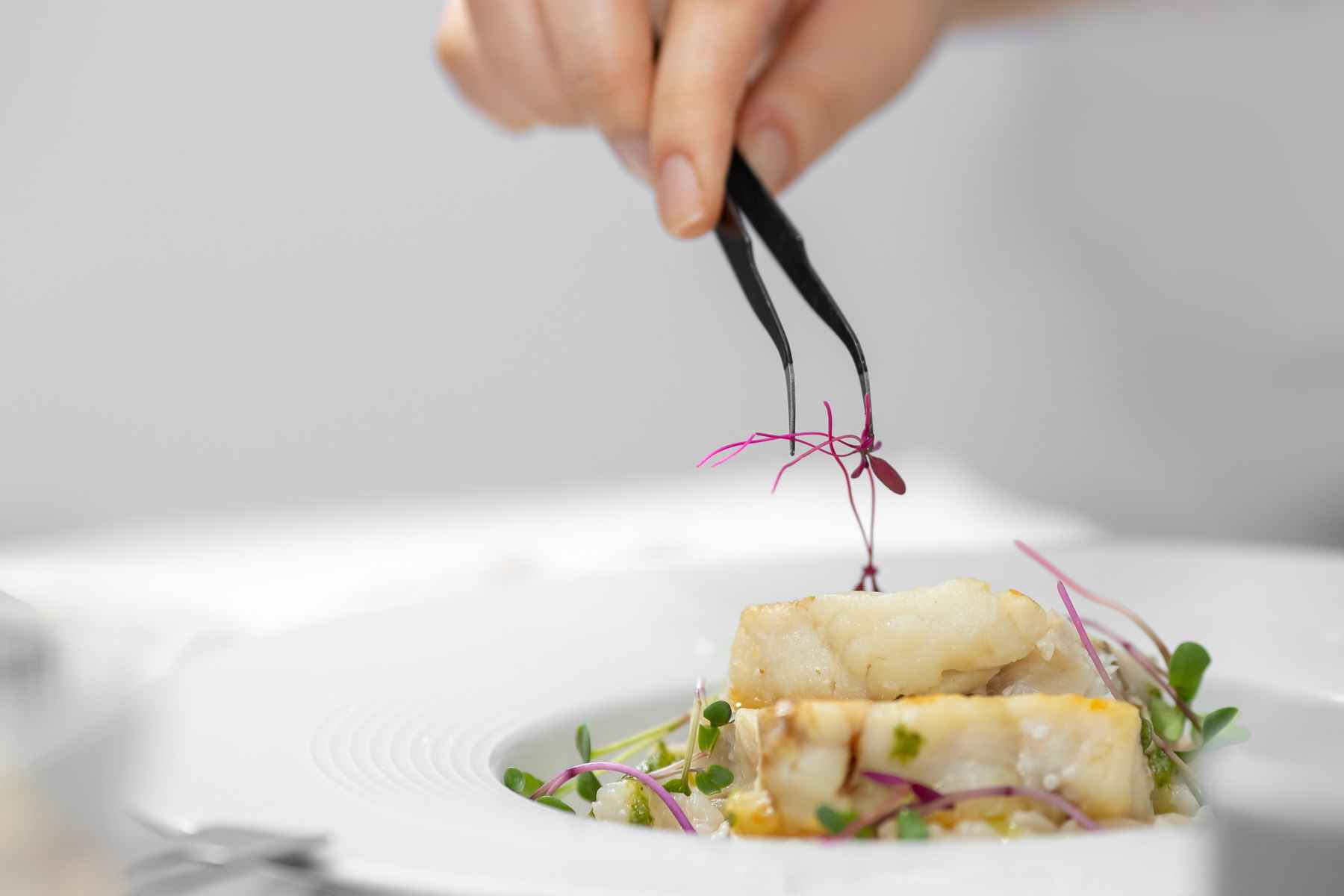Closeup of chef's hand using tweezers to plate micro herbs on a dinner plate. Favorite kitchen tools.