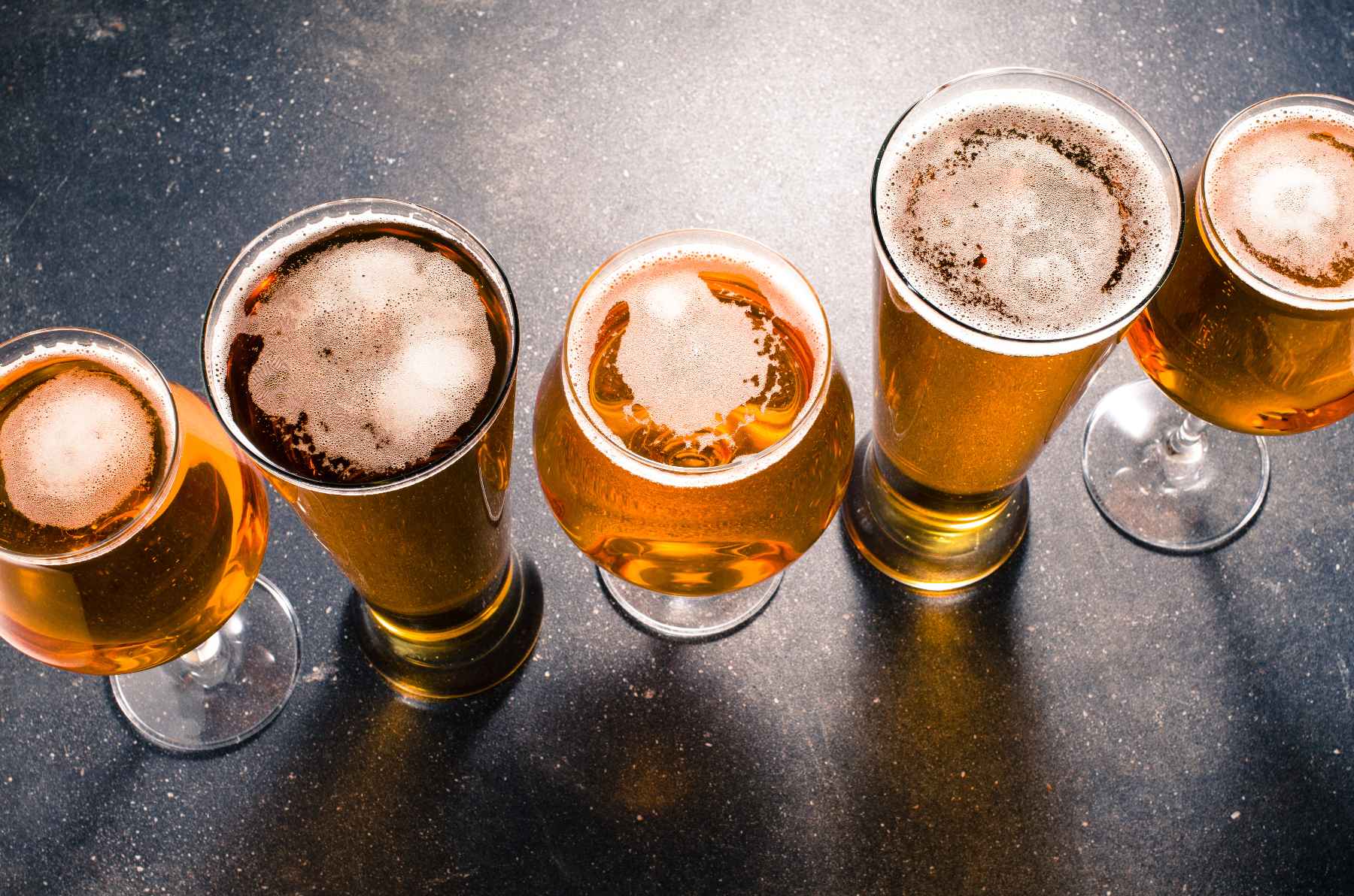 Photo of five amber beers in pint and tulip glasses lined up on a black slate counter. Getting a Colorado liquor license.