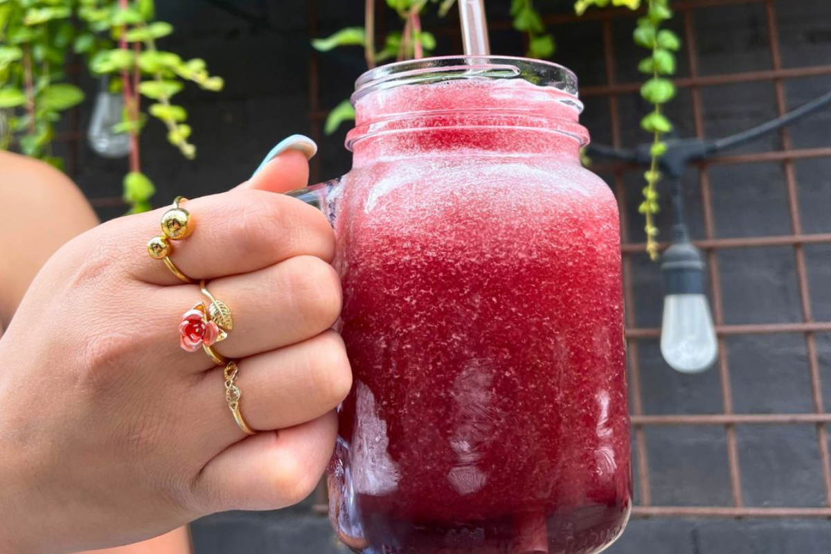 Acova_The Misty 2_Photo Courtesy of Acova A close-up of Acova’s Misty, a frozen red cocktail served in a mason jar, held by a hand wearing gold rings with a plant-filled patio in the background.