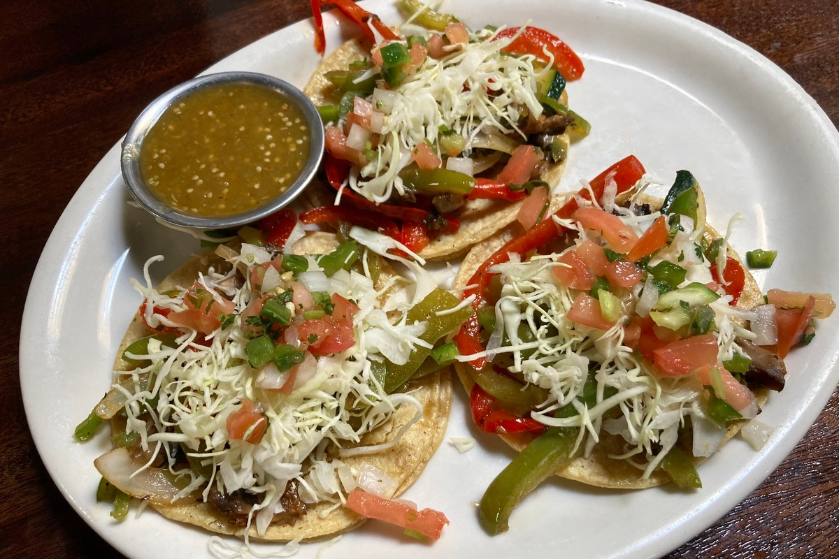 Adelitas_Tacos_by_Abigail_Bliss Three tacos from Adelitas topped with sautéed peppers, onions, shredded cabbage, and fresh pico de gallo, served with a side of green salsa on a white plate.