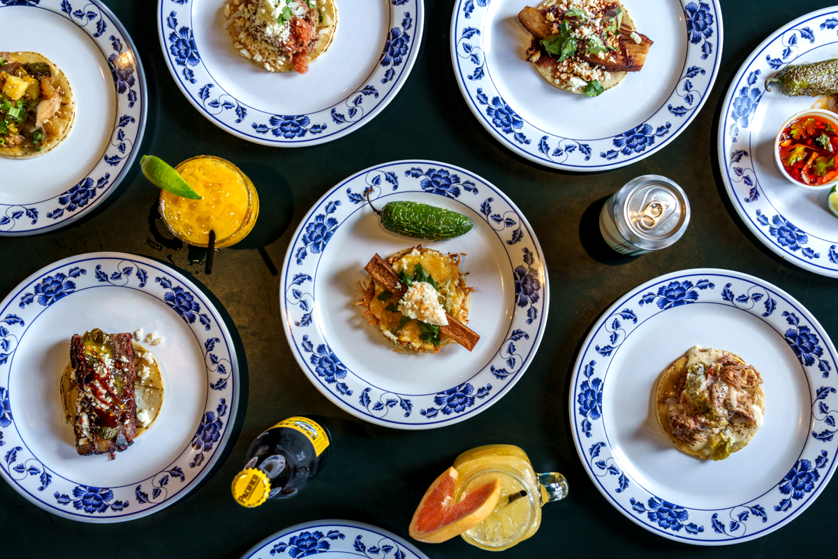 Adobo_by_Ashley_Beguin An overhead view of Adobo’s colorful taco spread, each plated on floral china, paired with cocktails, soda, and garnished with fresh lime and jalapeños.