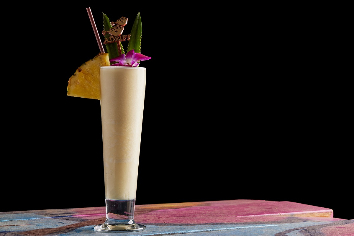 Adrift Chi Chi_Photo Credit_Harrison Warters Photography Adrift’s Chi Chi cocktail served in a tall glass with pineapple, orchid, and palm leaf garnish on a colorful wood table with a black background.