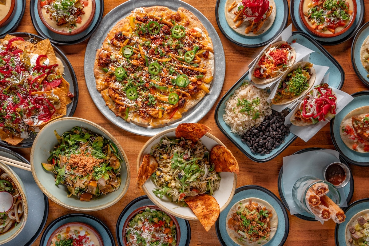 Los_Chingones_by_TAG_Restaurant_Group Colorful spread from Los Chingones featuring tacos, nachos, flatbread, rice and beans, salads, queso dips, and churros served on a wood table.