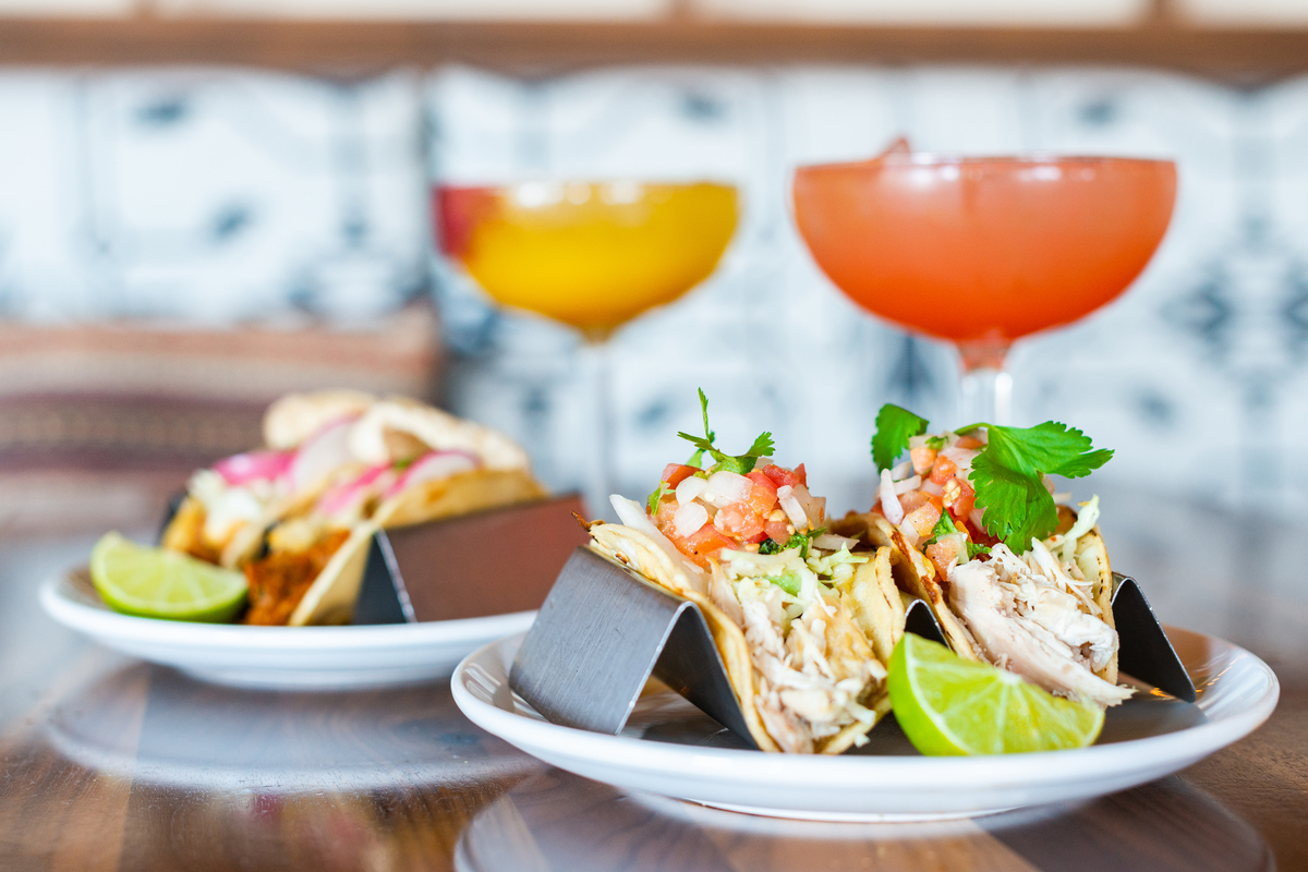 My_Neighbor_Felix_Tacos Two plates of tacos from My Neighbor Felix are served with lime wedges and topped with shredded chicken, pico de gallo, and cilantro, with cocktails in the background.