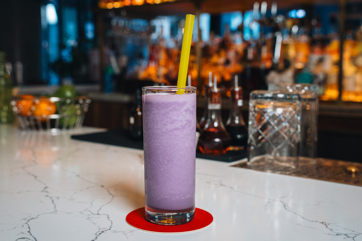 Poka Lola Social Club_Bad News Bears_Photo Courtesy of Poka Lola Social Club A tall glass of Poka Lola Social Club’s Bad News Bears slushy cocktail, bright purple in color with a yellow straw, served on a white marble bar.