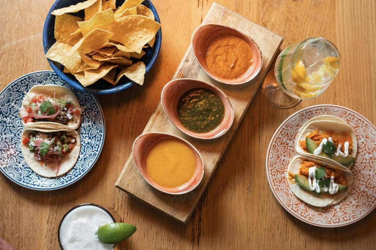 Taco Tuesday spread from Kachina Cantina with chips, tacos, and three kinds of salsa served on colorful plates with beer and a citrus cocktail on the table.