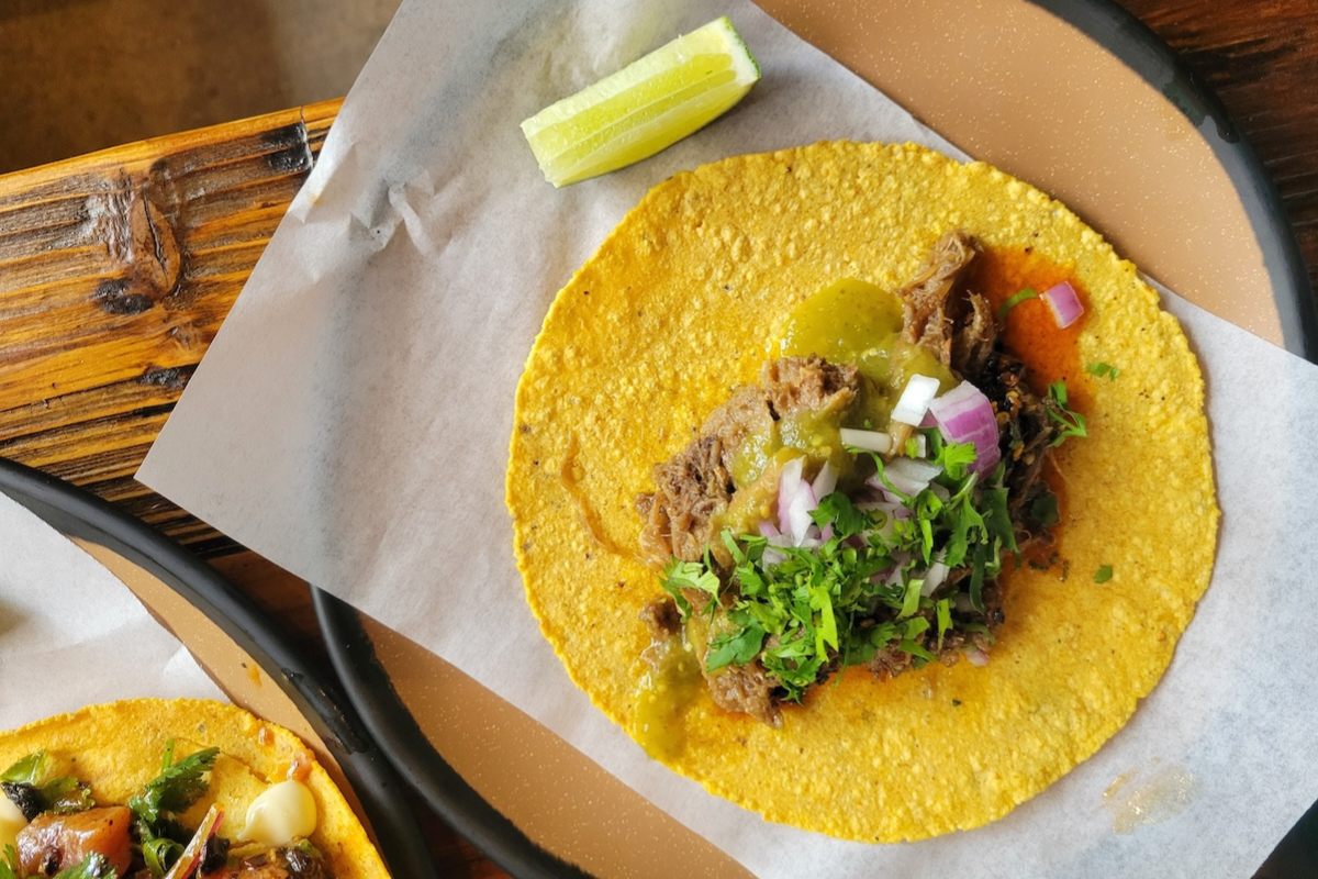 Xiquita_Lengua_Taco_by_Linnea_Covington A lengua taco from Xiquita topped with chopped onions, fresh cilantro, and green salsa on a yellow corn tortilla, served with a lime wedge on the side.