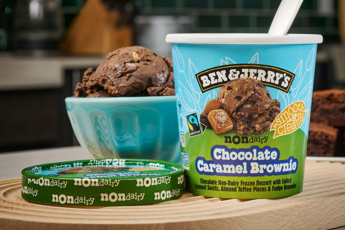 ben jerrys ice cream Ben & Jerry’s non-dairy Chocolate Caramel Brownie ice cream pint next to a bowl filled with scoops of the same flavor, sitting on a wooden tray.
