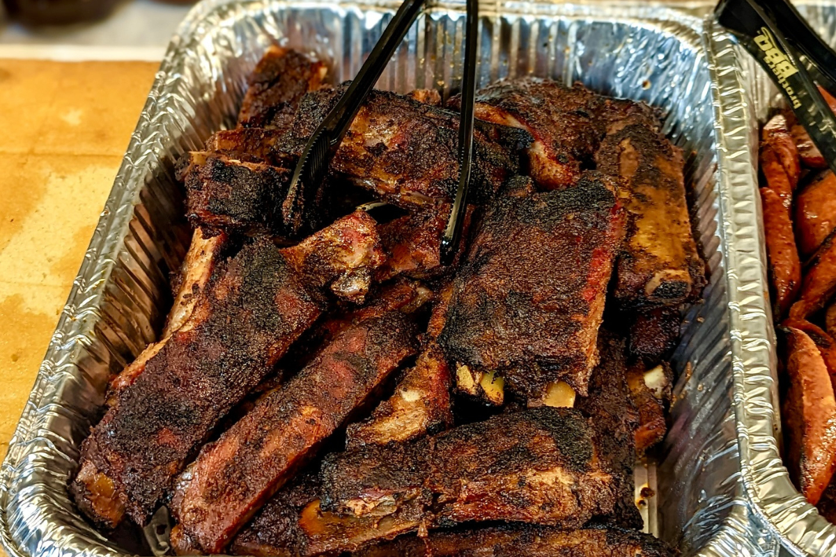 brothers bbq Tray of smoked ribs from Brothers BBQ, piled high in a foil pan with tongs ready to serve. Dark bark and smoky edges show off a slow-cooked finish.