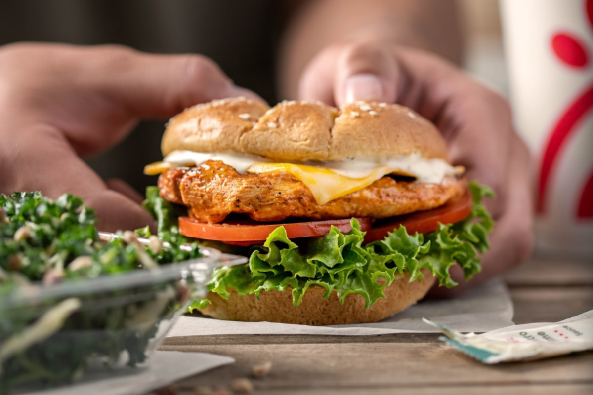 chick fil a Chick-fil-A grilled chicken sandwich with lettuce, tomato, cheese, and sauce on a wheat bun, served with a side salad and a drink cup in the background.