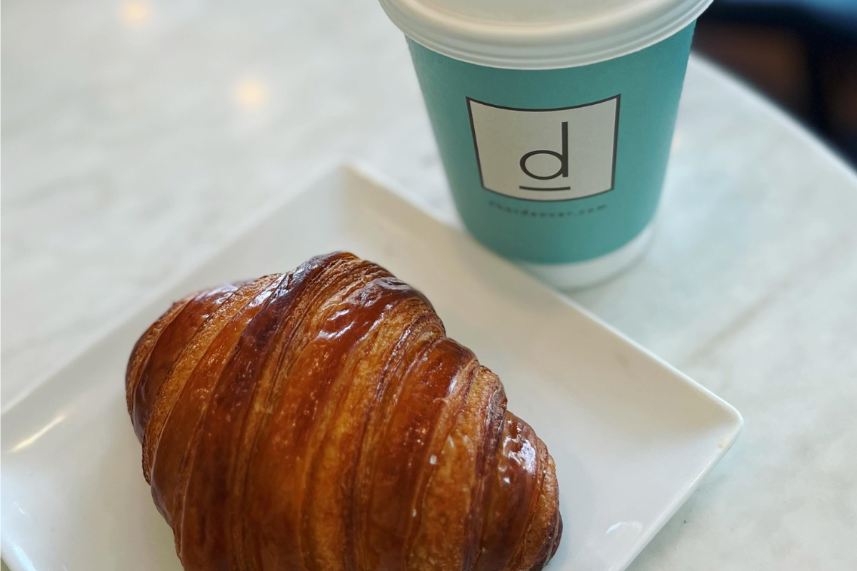 d bar A flaky, golden croissant on a white plate next to a blue takeaway coffee cup with the D Bar logo, set on a marble table.
