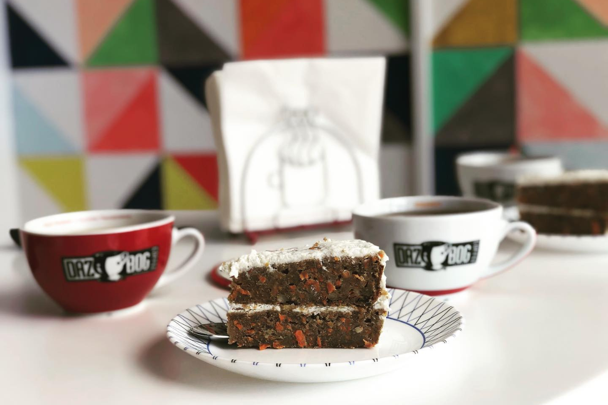 dazbog coffee A slice of carrot cake with white frosting on a plate, served with Dazbog coffee in branded mugs, set against a colorful geometric wall.