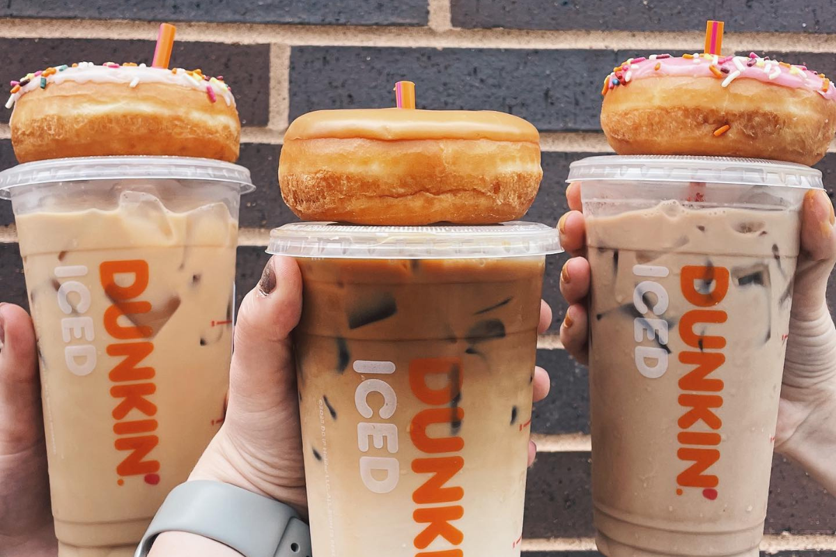 dunkin Three hands holding Dunkin' iced coffee drinks with donuts placed on top of each lid, two with sprinkles and one glazed, against a brick wall background.
