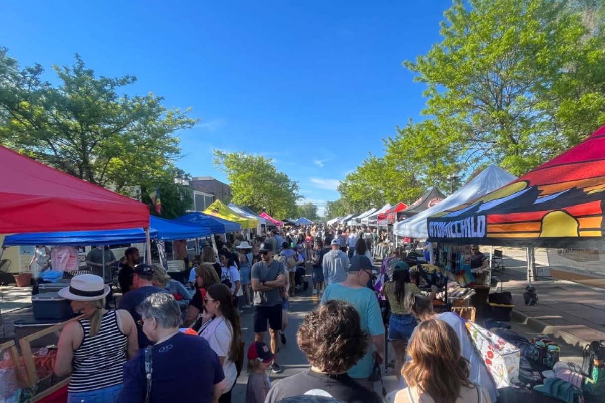 erie farmers market Shoppers walk through Erie Farmers Market on a sunny day, surrounded by colorful tents, green trees, and vendors selling handmade goods, produce, and treats.