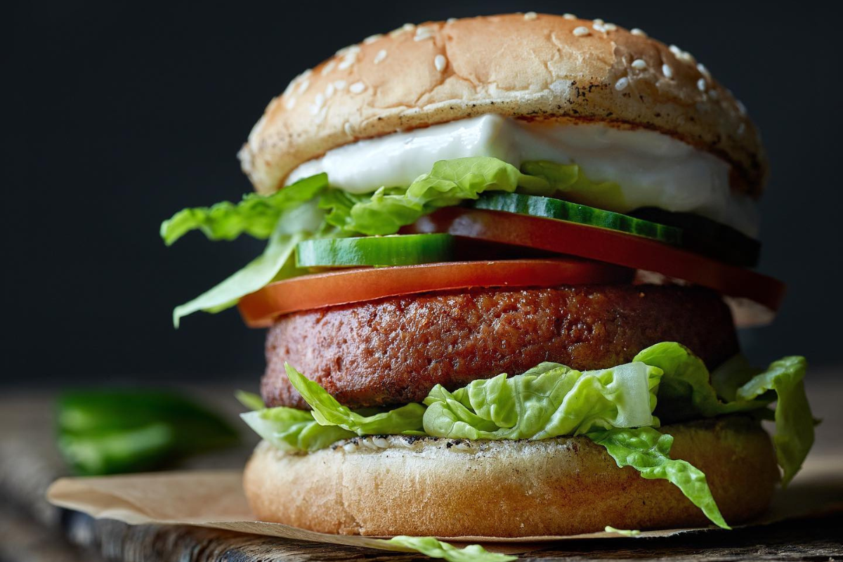 etais cafe bar A close-up of a veggie burger stacked with lettuce, tomato, cucumber, and a sesame seed bun, served on parchment paper with a dark background.