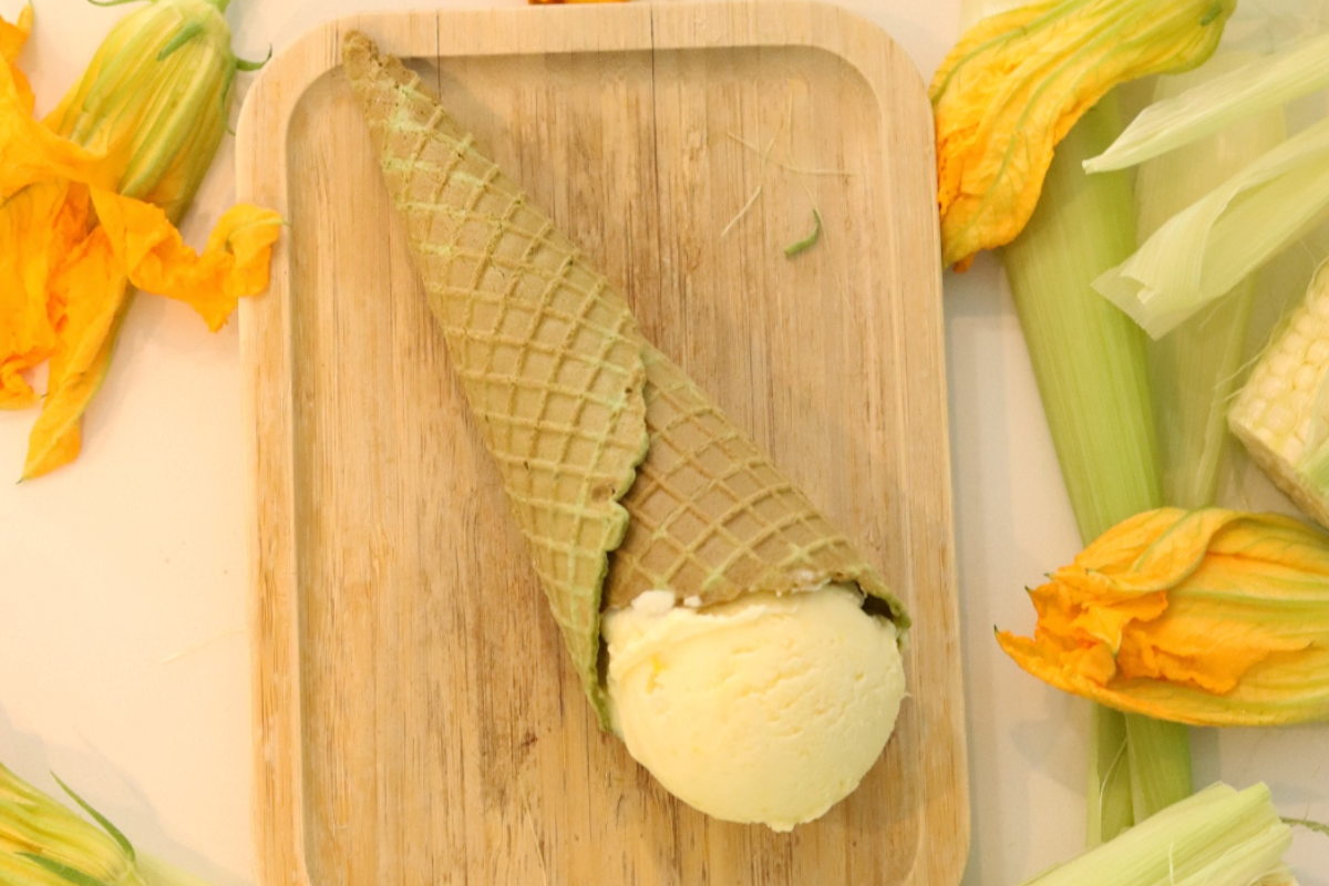 heaven creamery Corn bread gelato scoop in a green waffle cone on a wooden tray, surrounded by fresh corn husks and squash blossoms.