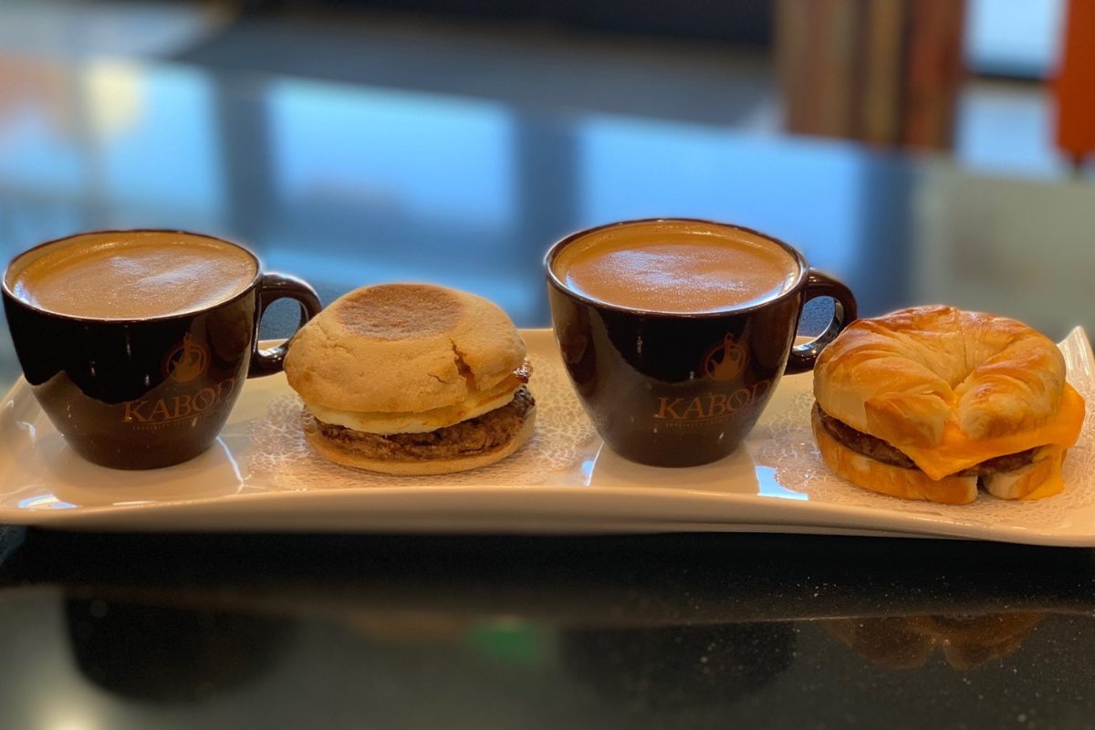 kabod coffee Two Kabod coffee mugs with frothy drinks served alongside a sausage English muffin and a croissant breakfast sandwich on a white tray.