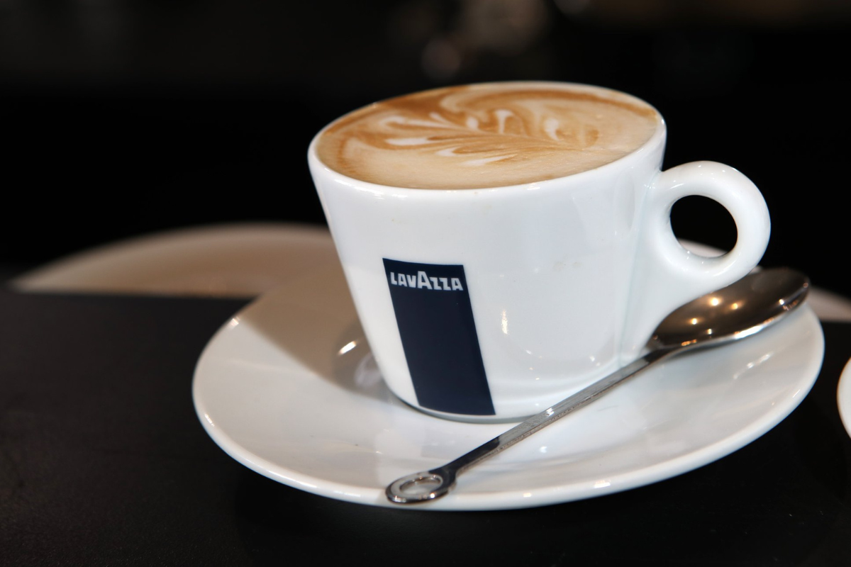 lavazza A Lavazza-branded white cup filled with latte art on top, placed on a matching saucer with a silver spoon, set against a dark background.