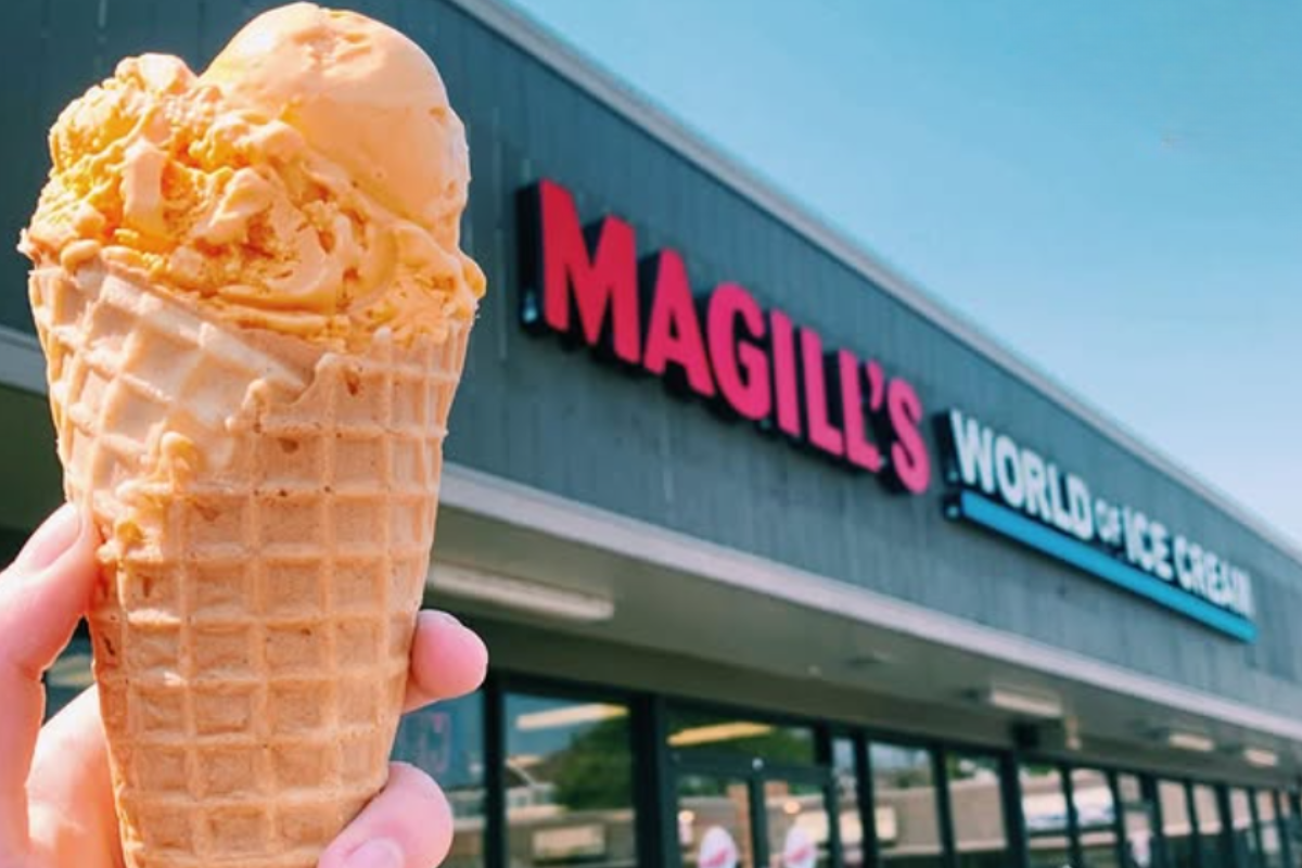 magills world of ice cream Close-up of an orange ice cream cone held in front of a storefront with a bright sky, showing creamy texture and crunchy waffle cone details.