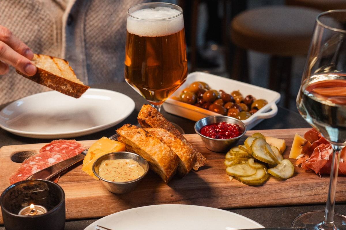 mercantile dining and provision Charcuterie board with sliced meats, cheeses, pickles, bread, and spreads served with a glass of beer and white wine at Mercantile Dining & Provision.