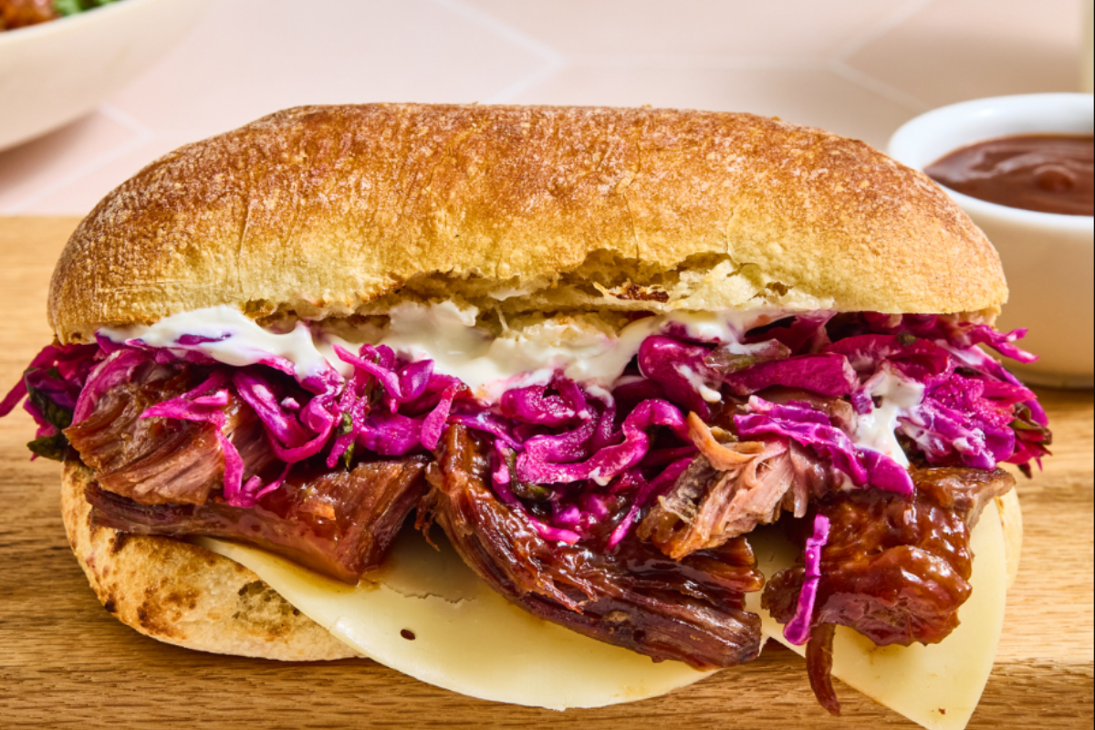 modern market eatery Close-up of a pulled beef sandwich with melted cheese, creamy sauce, and vibrant purple slaw on toasted ciabatta bread, served with a side of BBQ sauce.