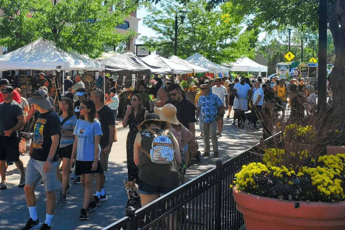 parker farmers market Shoppers walk through the busy Parker Farmers Market on a sunny day, browsing vendor tents filled with food, flowers, and handmade goods under shady trees.