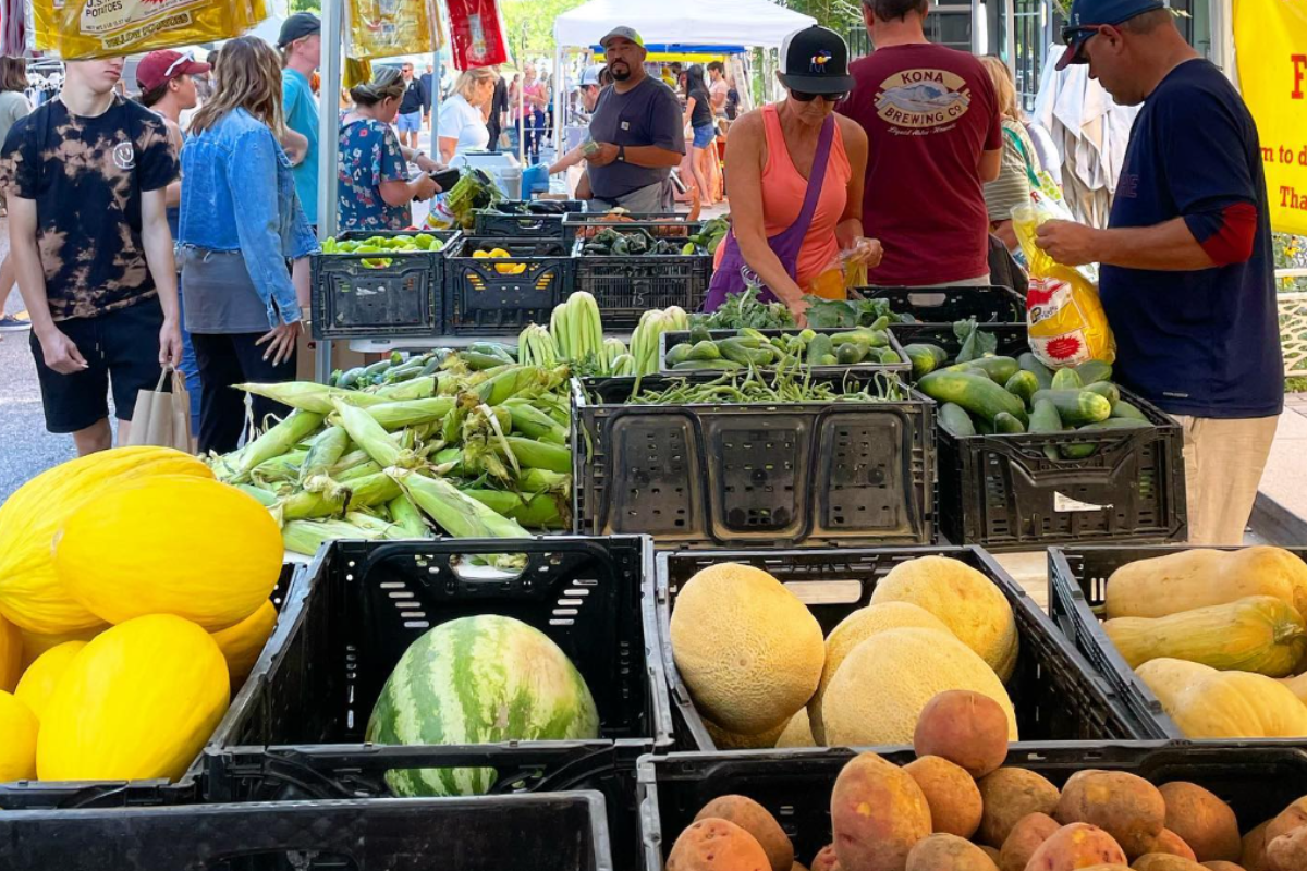 people produce at belleview station People + Produce at Belleview Station in Denver with shoppers browsing fresh produce like melons, corn, cucumbers, and potatoes under shaded vendor tents.