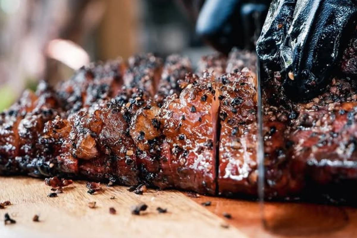 piggin out smokehouse Close-up of sliced brisket from Piggin’ Out Smokehouse, glistening with fat and cracked pepper, being cut on a wooden board by a gloved hand.
