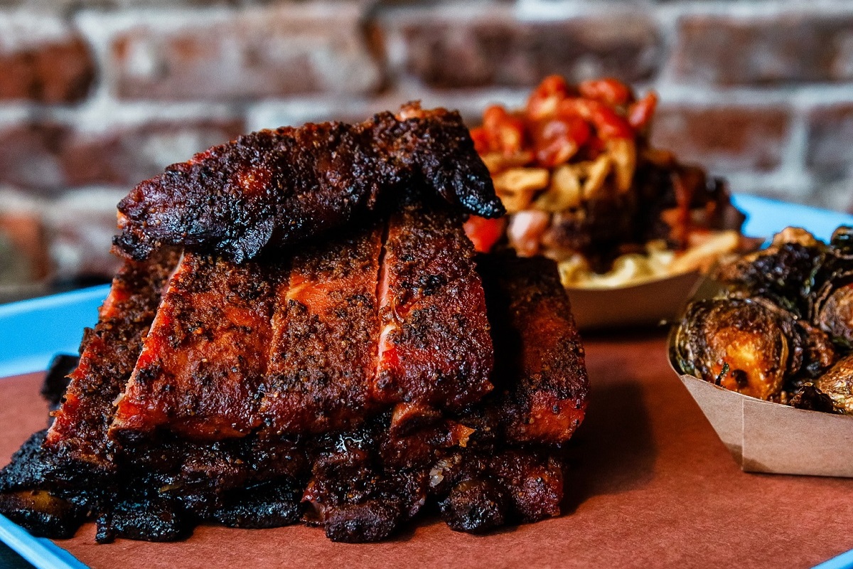 post oak barbecue Stack of smoked pork ribs with a dark bark and red smoke ring, served on a tray with crispy Brussels sprouts and loaded fries in the background.
