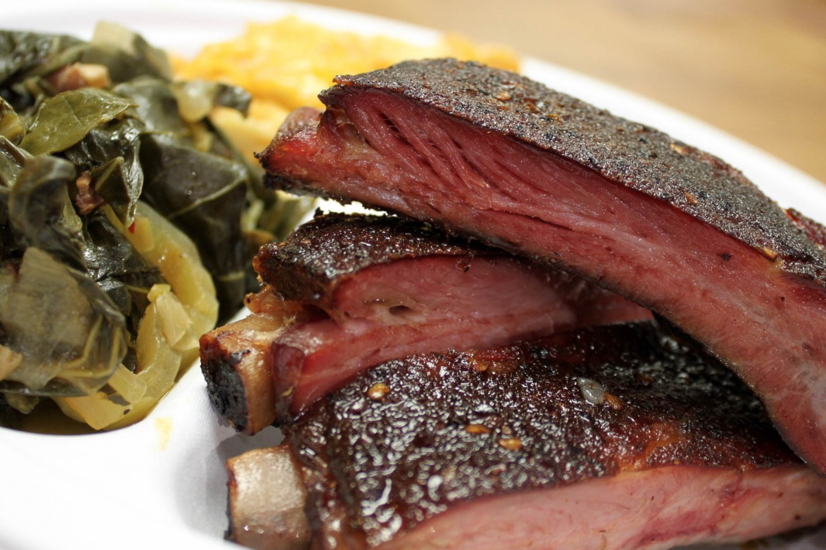 rolling smoke bbq Close-up of smoked pork ribs from Rolling Smoke BBQ, served with tender collard greens and creamy mac and cheese on a white plate.