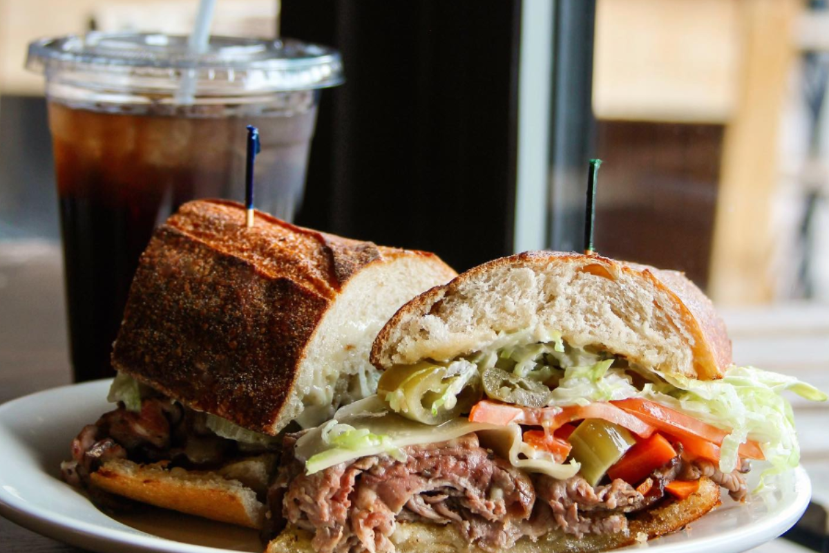 salt grinder Salt & Grinder sandwich stacked with roast beef, shredded lettuce, tomatoes, peppers, and cheese on crusty bread, served with iced soda in the background.