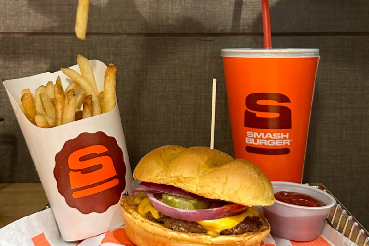 smashburger bar Smashburger meal with cheeseburger, fries in branded cup, ketchup, and a drink in an orange cup with logo on a tray against a brown background.