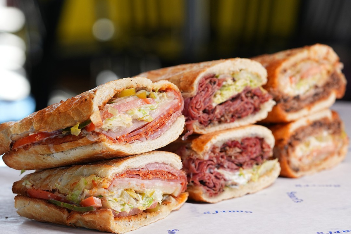 snarfs sandwiches Three types of toasted sub sandwiches from Snarf’s, each cut in half and stacked, filled with meats, cheese, lettuce, tomatoes, and pickles.