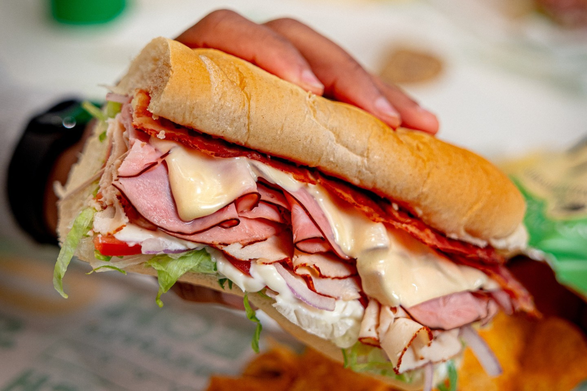 subway A hand holding a Subway sandwich stacked with deli meats, melted cheese, lettuce, tomato, onion, and mayo in a fresh sub roll.