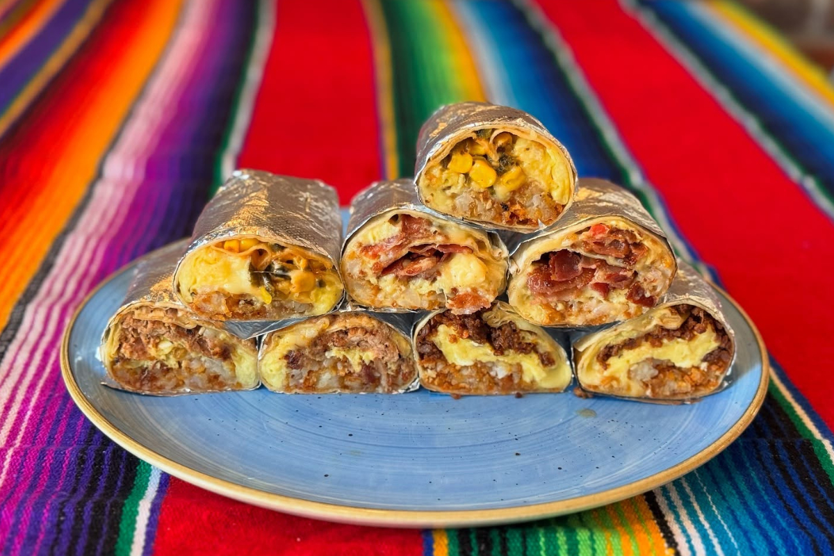 tacos tequila whiskey A plate stacked with foil-wrapped breakfast burritos, sliced to show fillings like eggs, meat, cheese, and veggies, on a colorful Mexican-style tablecloth.
