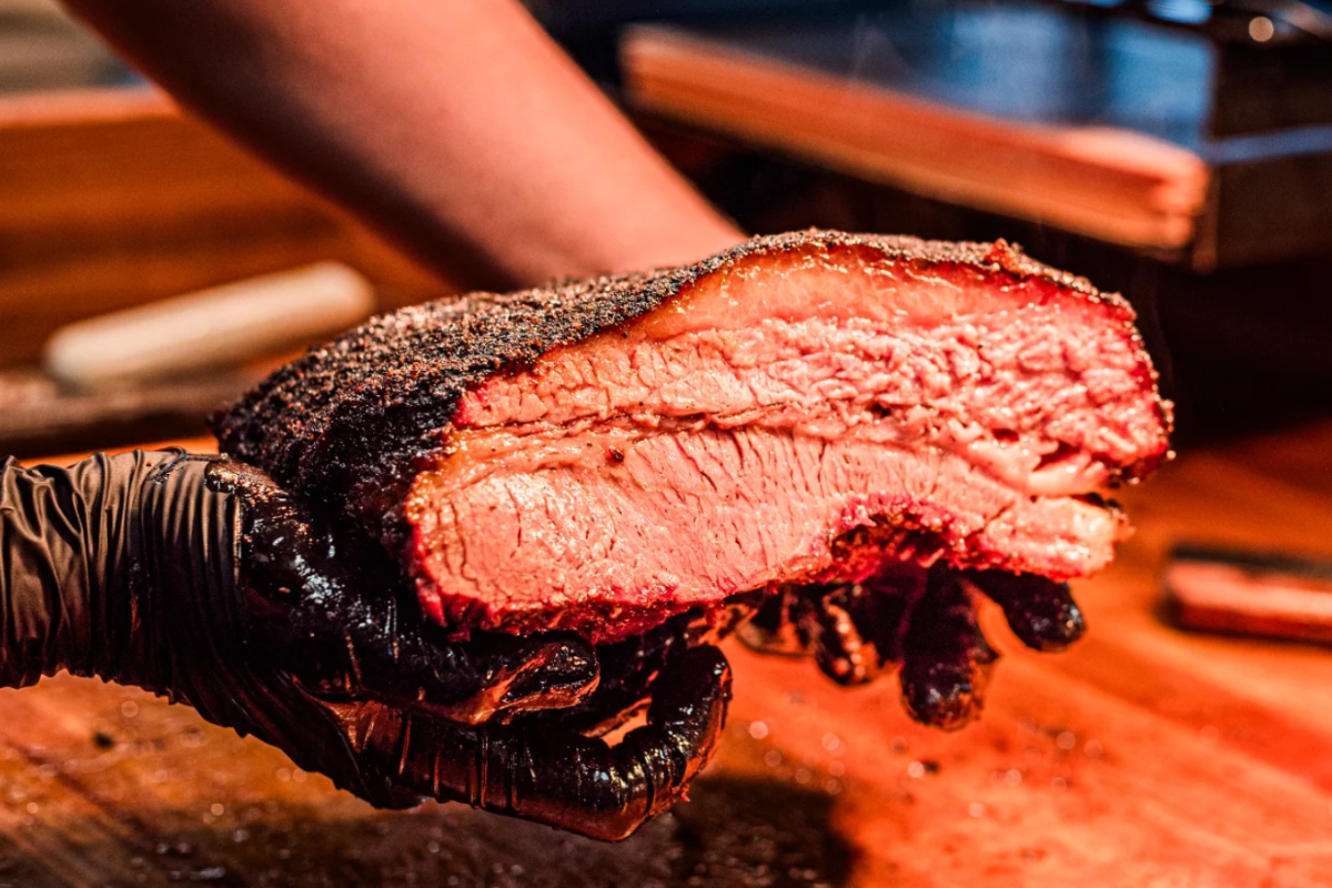 waynes smoke shack Close-up of a juicy smoked brisket slice from Wayne's Smoke Shack, held in gloved hands, showing its bark, fat cap, and pink smoke ring.