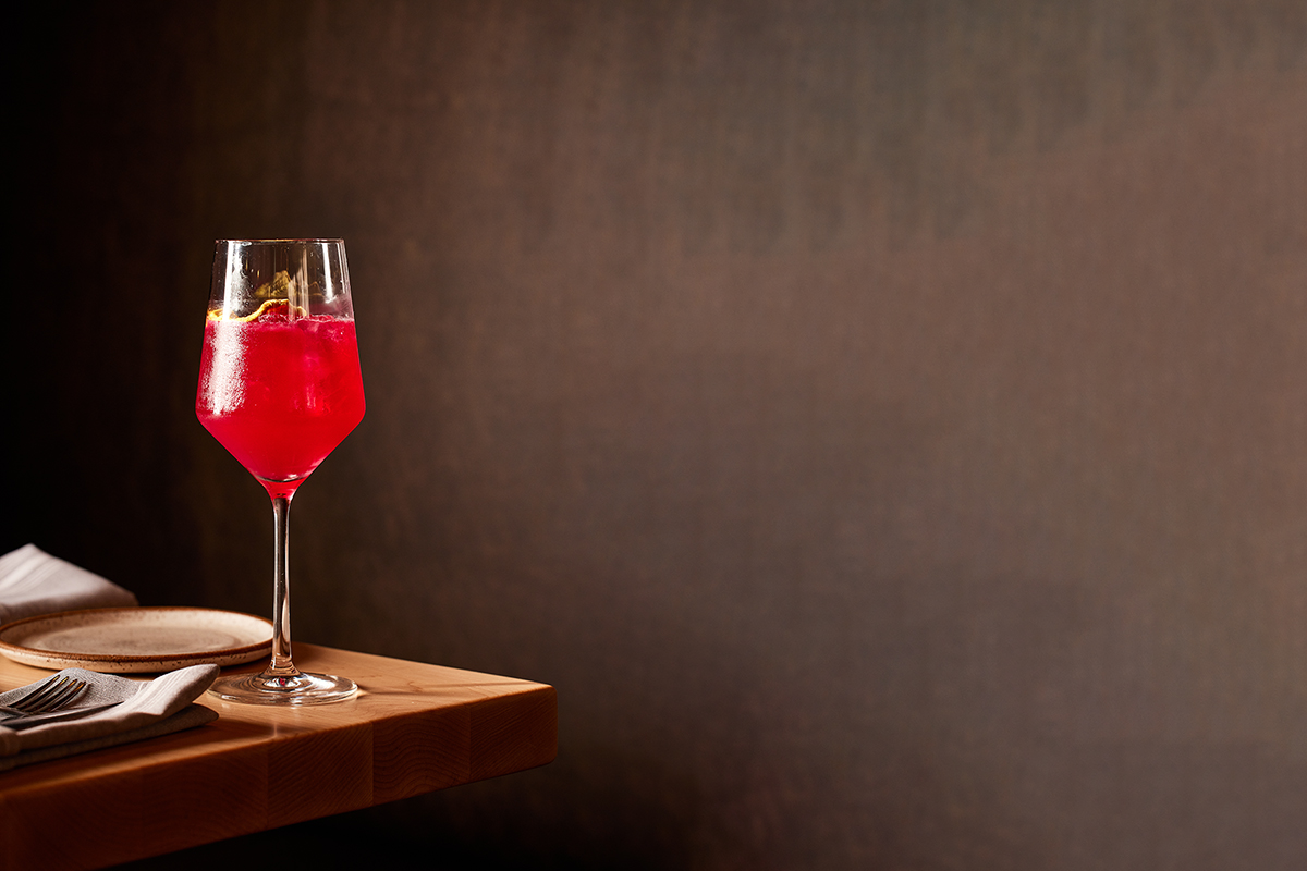 Bright pink beet spritz cocktail served in a tall wine glass with ice and lemon twist, placed on a wooden table with napkin and plate.