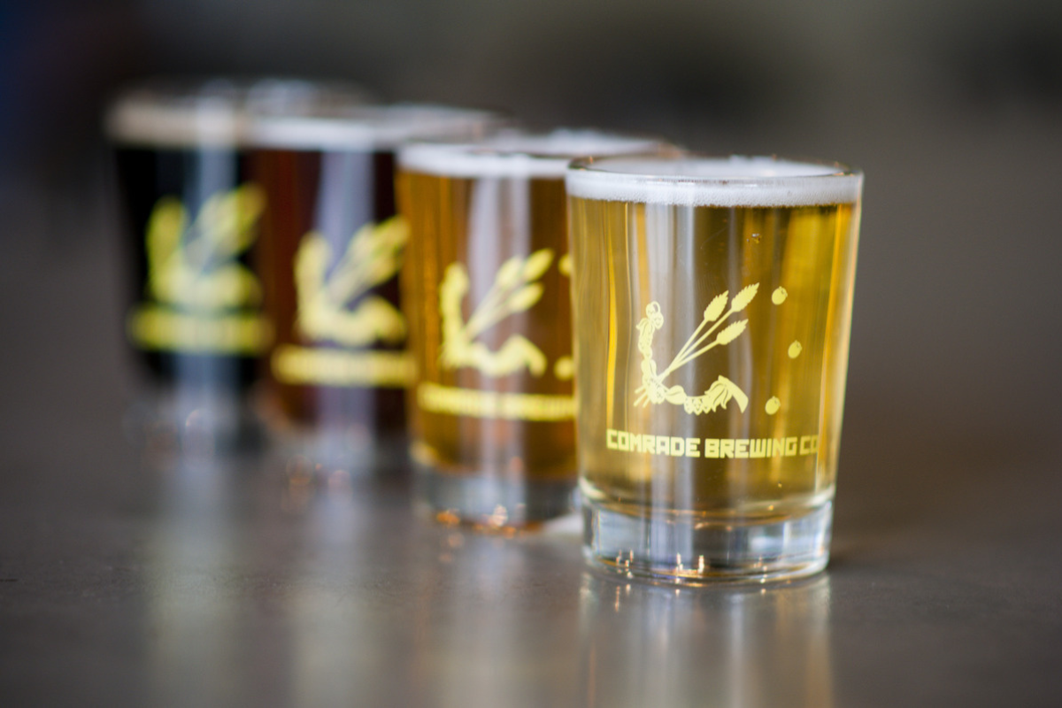 Comrade_Brewing_Company Flight of beers from Comrade Brewing Company in Denver, Colorado, featuring a range of styles in branded tasting glasses lined up on a bar counter.