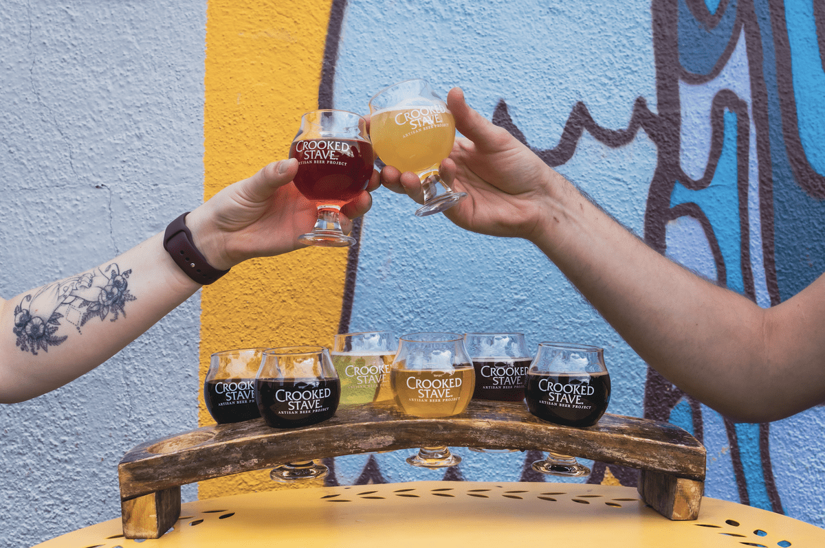 Crooked_Stave_Brewing_Company Two people cheers with Crooked Stave beers over a colorful wall, with a rustic beer flight tray featuring various artisan brews on a yellow table.