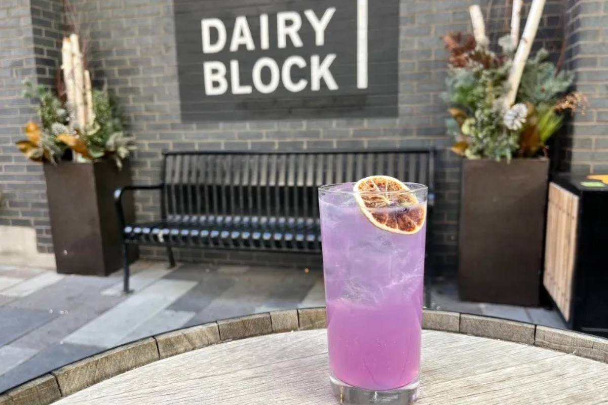 Purple Rockies Fizz cocktail topped with a dried citrus slice, served over ice in a tall glass on a patio table in front of Dairy Block’s brick wall sign.