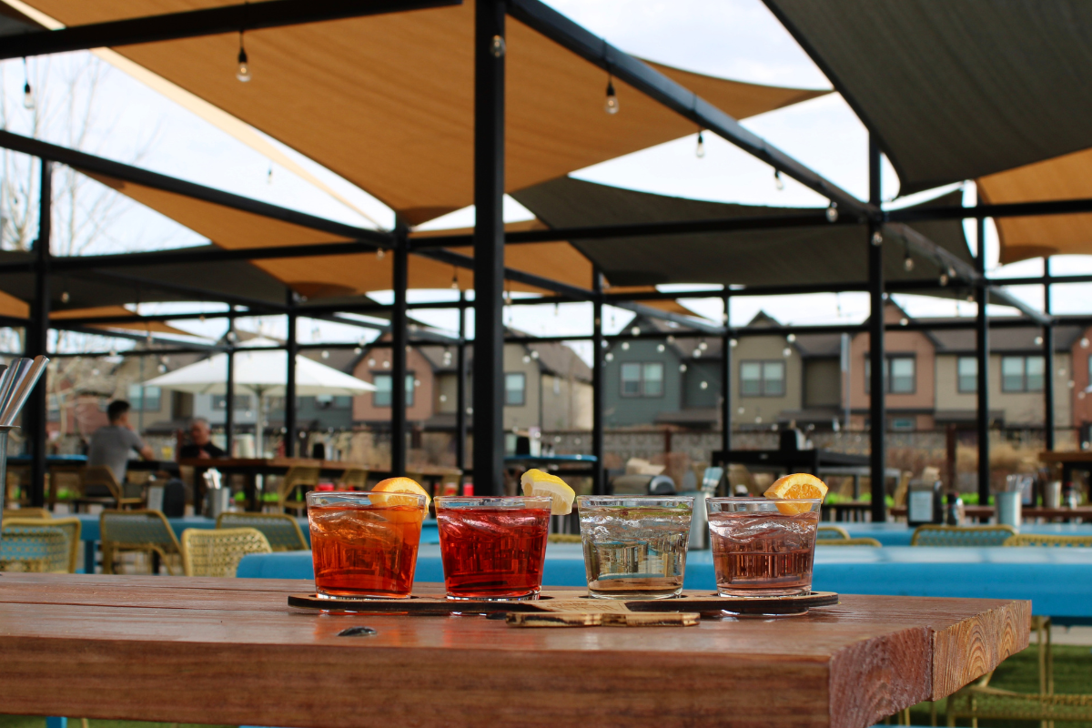 Spritz flight with four colorful cocktails garnished with citrus, served on a wood table at FlyteCo Tower’s outdoor patio with shaded seating.