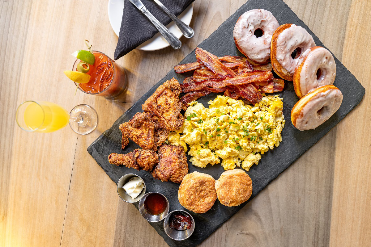 Holy $ht Breakfast_photo courtesy of Church Union A slate serving board loaded with scrambled eggs, crispy bacon, fried chicken, buttermilk biscuits, and glazed donuts, with a Bloody Mary and mimosa on the side.