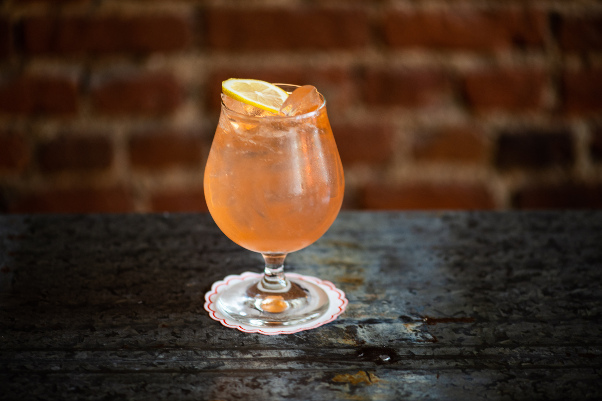 Casoni Spritz served over ice in a stemmed glass with a lemon slice at Jovanina’s, placed on a metal table with a brick wall in the background.