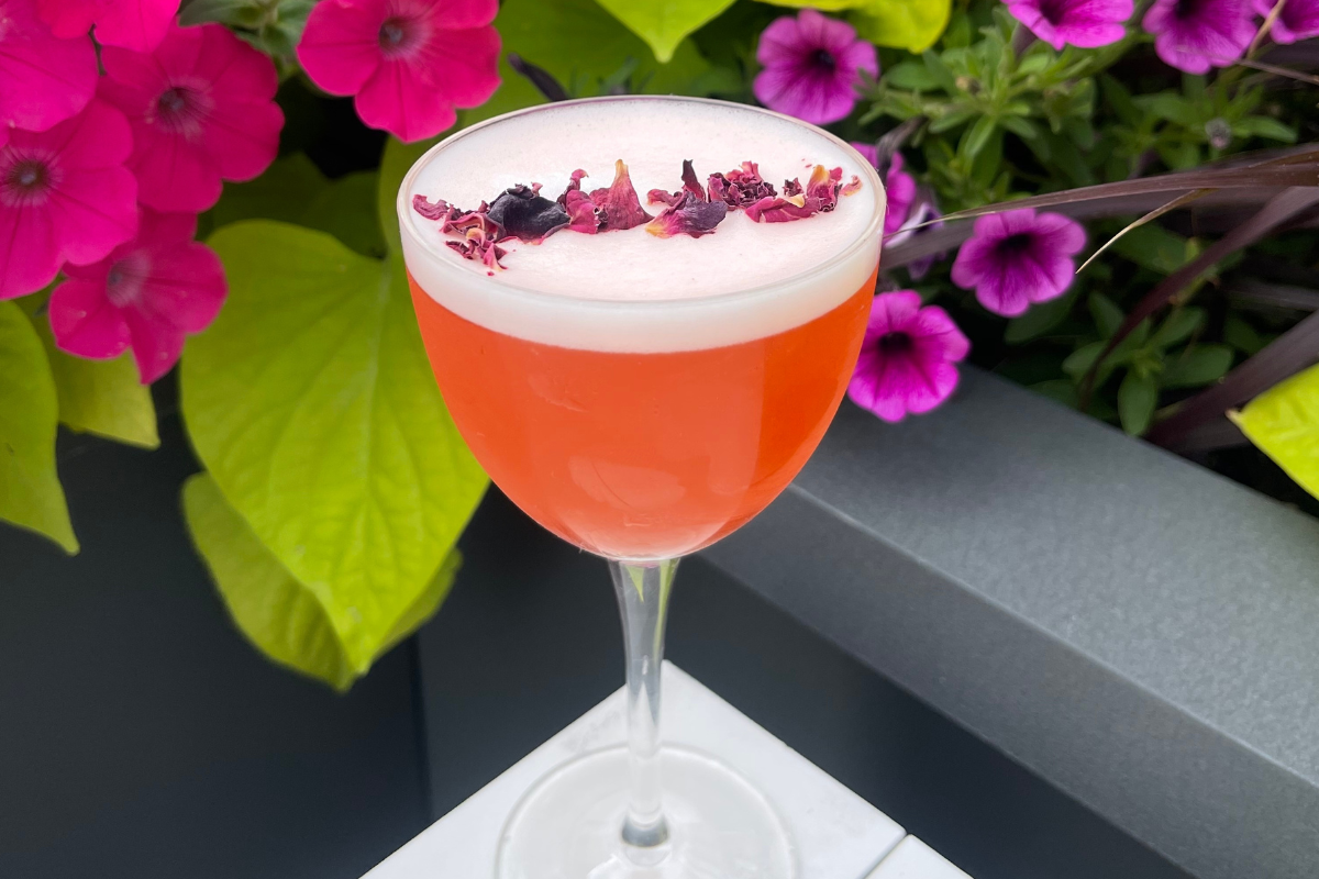 Strawberry Tides spritz with a foamy top and dried flower garnish, served in a coupe glass at Kisbee on the Roof with bright flowers in the background.
