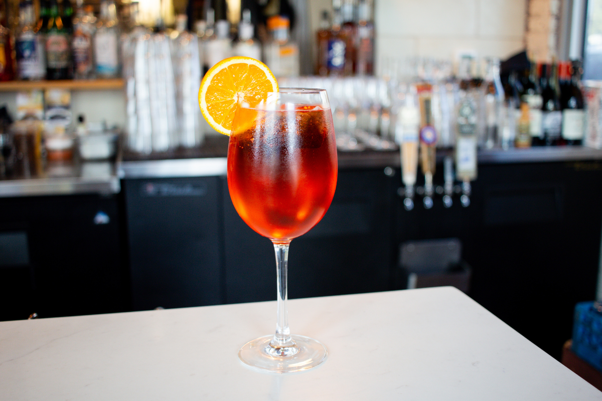 Leven Deli Co.’s house spritz served in a wine glass with ice and an orange slice garnish, set on a white counter with a bar backdrop.