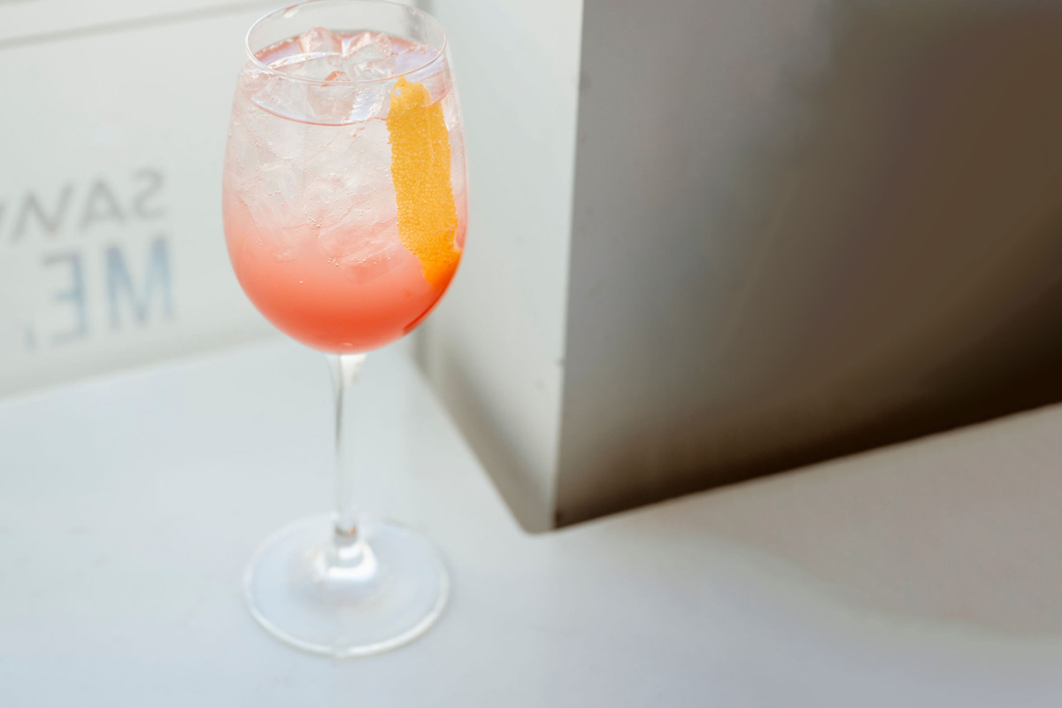 Barcelona Spritz from Olive & Finch, a light pink cocktail with ice and an orange peel garnish, served in a wine glass on a sunlit white tabletop.