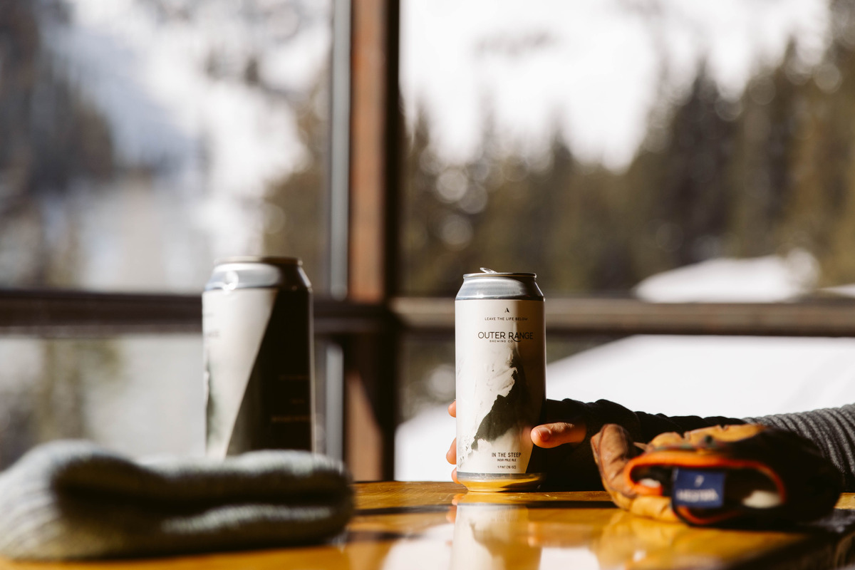 Outer_Range_Brewing_Company Can of Outer Range Brewing Company beer on a wooden table with ski gloves and beanie, set against a snowy mountain backdrop in Colorado.